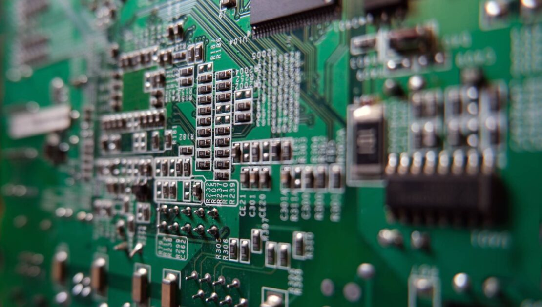 Close-up view of a green printed circuit board (PCB) showcasing surface mount technology (SMT) components such as resistors, capacitors, and integrated circuits, with visible traces and solder joints.