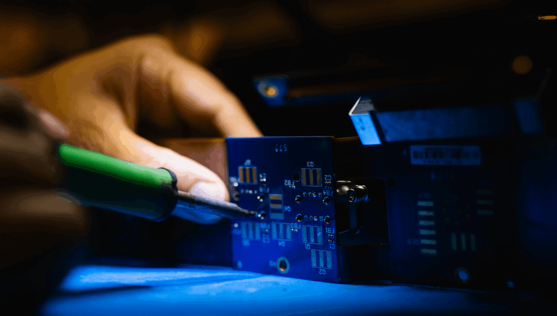 Close-up of a person soldering components onto a blue printed circuit board (PCB) during the electronics manufacturing process.