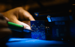 Close-up of a person soldering components onto a blue printed circuit board (PCB) during the electronics manufacturing process.