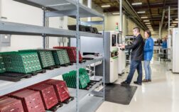 Outsourced electronics manufacturers operate automated equipment on an electronics manufacturing line beside racks of green and red printed circuit boards.