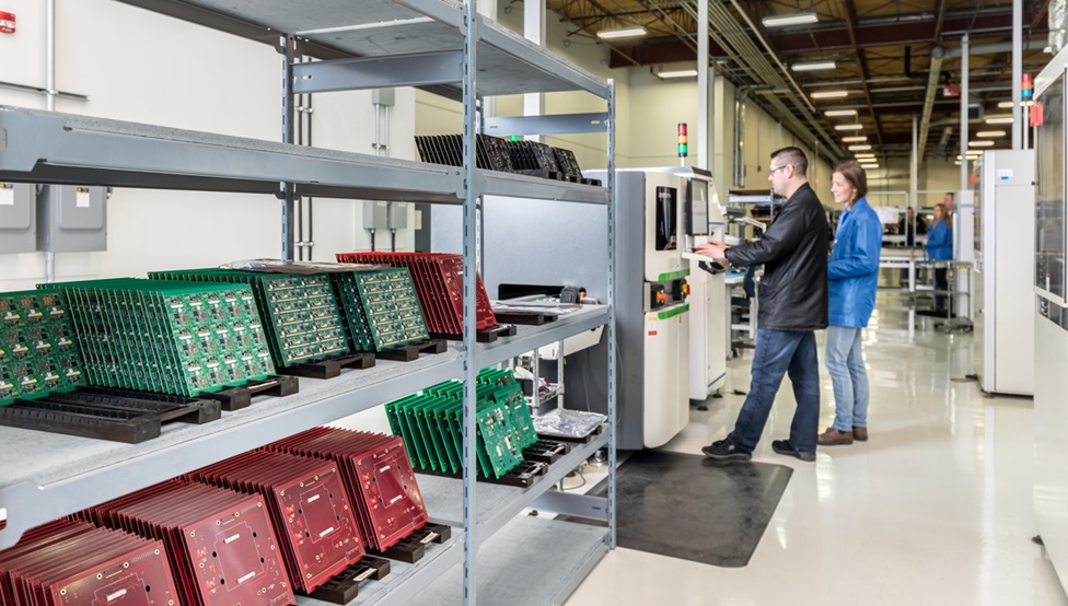 Outsourced electronics manufacturers operate automated equipment on an electronics manufacturing line beside racks of green and red printed circuit boards.