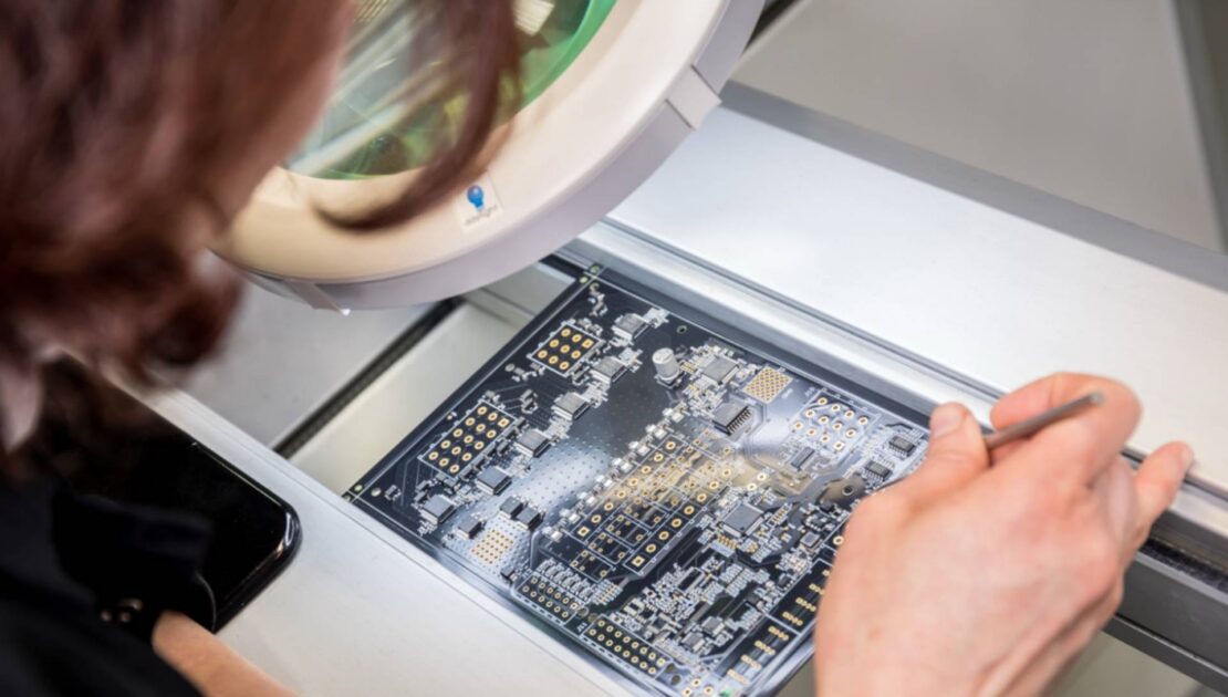 Technician inspecting a printed circuit board (PCB) under a magnifying lamp while holding precision tweezers at an electronics workstation.