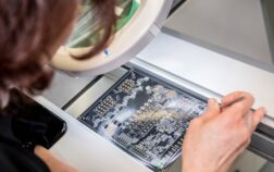 Technician inspecting a printed circuit board (PCB) under a magnifying lamp while holding precision tweezers at an electronics workstation.