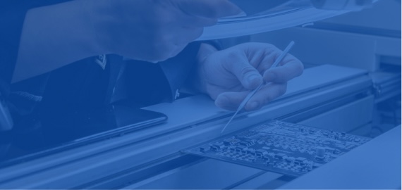 A hand holds a probe over a circuit board on an inspection line while another hand holds a document.